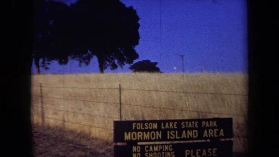 Visitor-shot 8mm view of the Mormon Island Area sign low in frame, with dry California grassland, fencing, and trees above it.