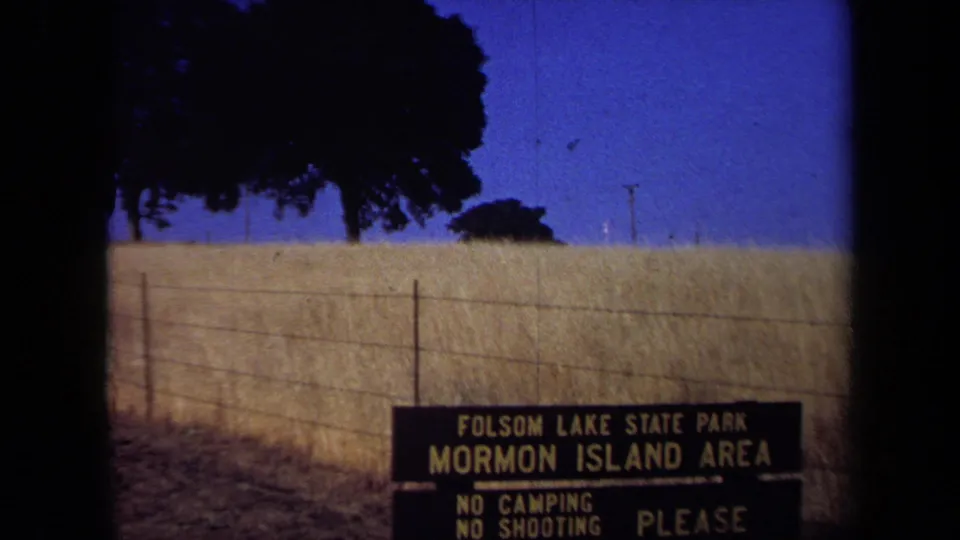 1971 California park sign for Folsom Lake State Park's Mormon Island Area, filmed with trees and fenced dry grassland behind it.