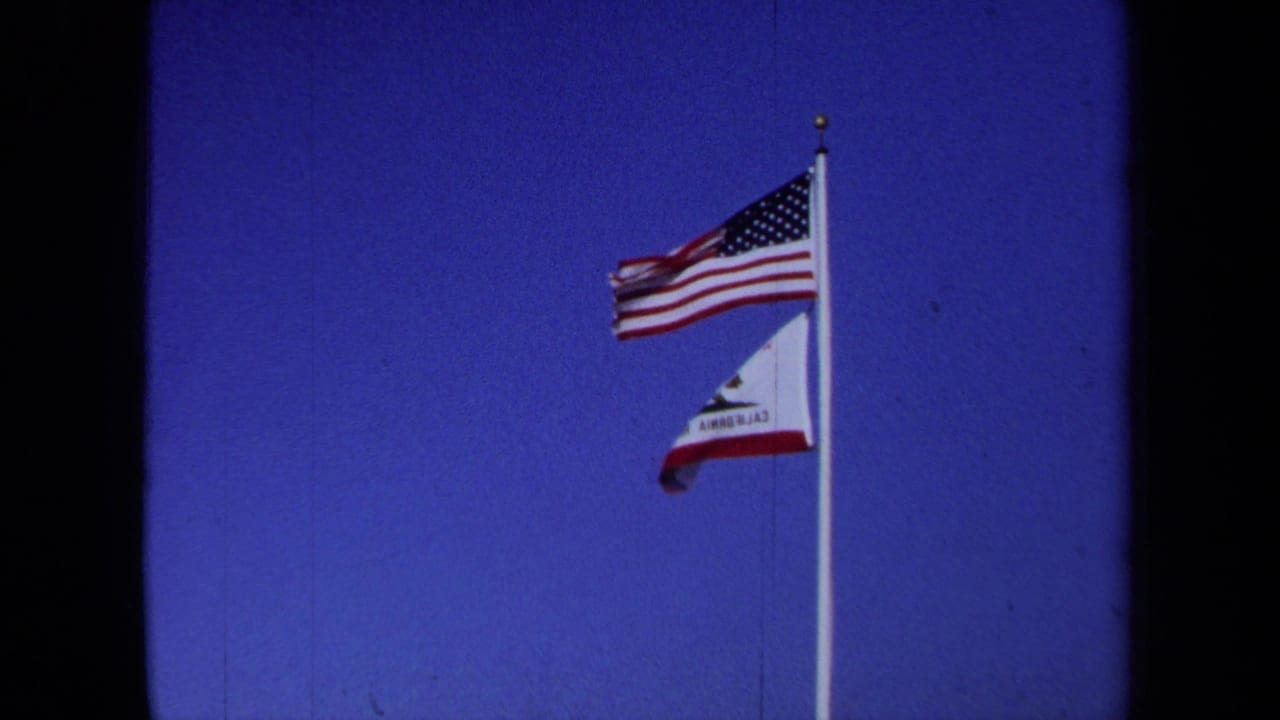 1971 California home-movie frame showing U.S. and California state flags waving together on a white flagpole.