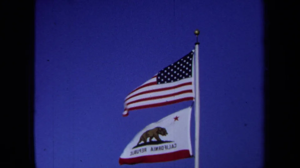 A closer 8mm frame shows the U.S. flag and California flag rippling in wind on the same pole.