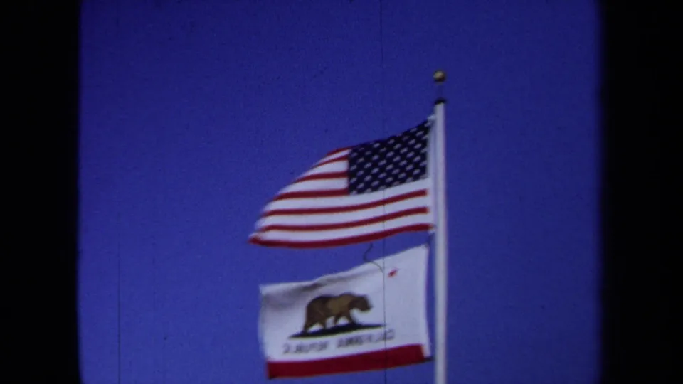 The final frame holds on the U.S. and California flags waving on a pole, with visible 8mm border and grain.