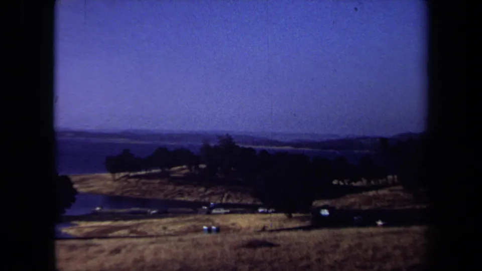 1971 California landscape frame with dry hills, dark tree lines, small pale details, and water on the horizon.