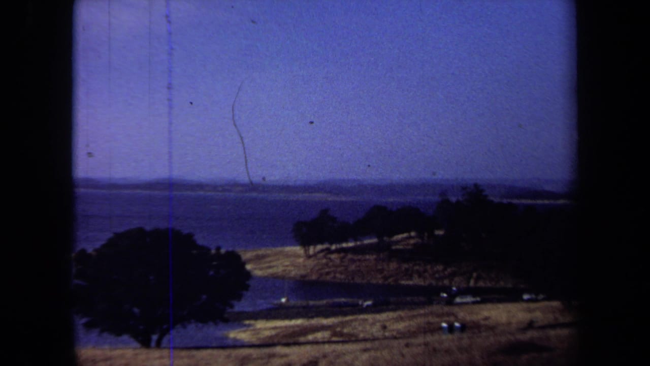 1971 California 8mm frame showing a lake bordered by dry grass, trees, and low hills under a clear sky.