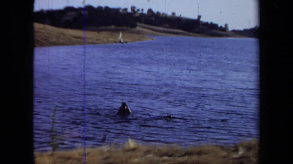 1971 California home-movie frame with a person swimming in rippled lake water near a dry shore and distant trees.