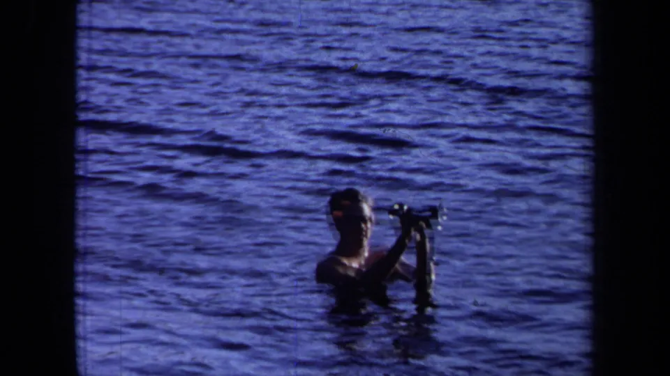 A man in open water holds a small camera or filming device up from the surface in this 1971 California 8mm frame.