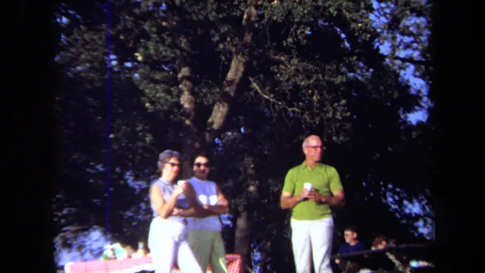 Close 8mm picnic frame of three adults in 1971 California, with cups and trees behind them.