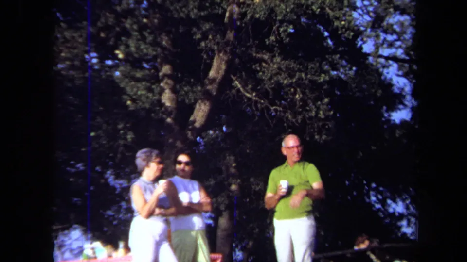 1971 California home-movie frame of adults holding cups near trees, with cars partly visible behind them.