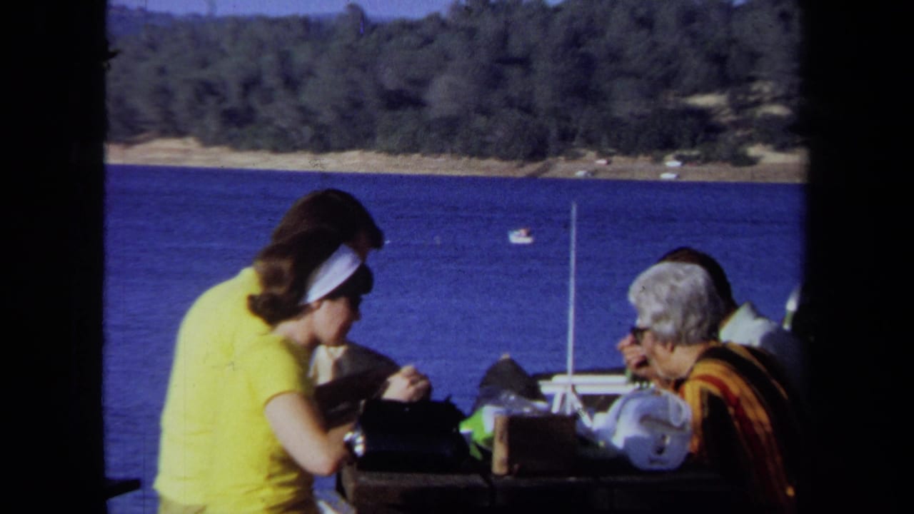 1971 California home-movie frame of family members eating at a lakeside picnic table with boats and forested shoreline behind them.