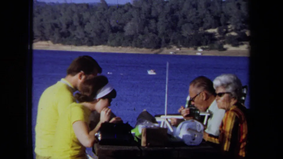 1971 California lakeside picnic scene with adults gathered around food and drink at a park table near blue water.