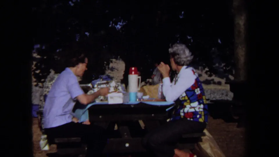 1971 California home-movie frame showing older adults eating at a shaded lakeside picnic table with a colorful vest and thermos visible.