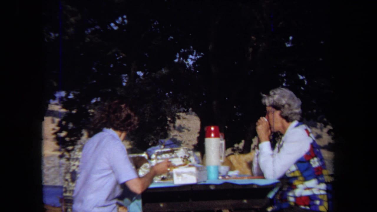In 1971 California home-movie footage, two women sit at a shaded picnic table with cups, food containers, and a red-capped thermos.