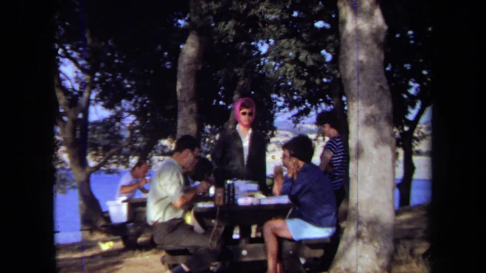 A wider 1971 California home-movie frame shows adults gathered at a lakeside picnic table, with trees, water, and a woman in a pink headscarf.