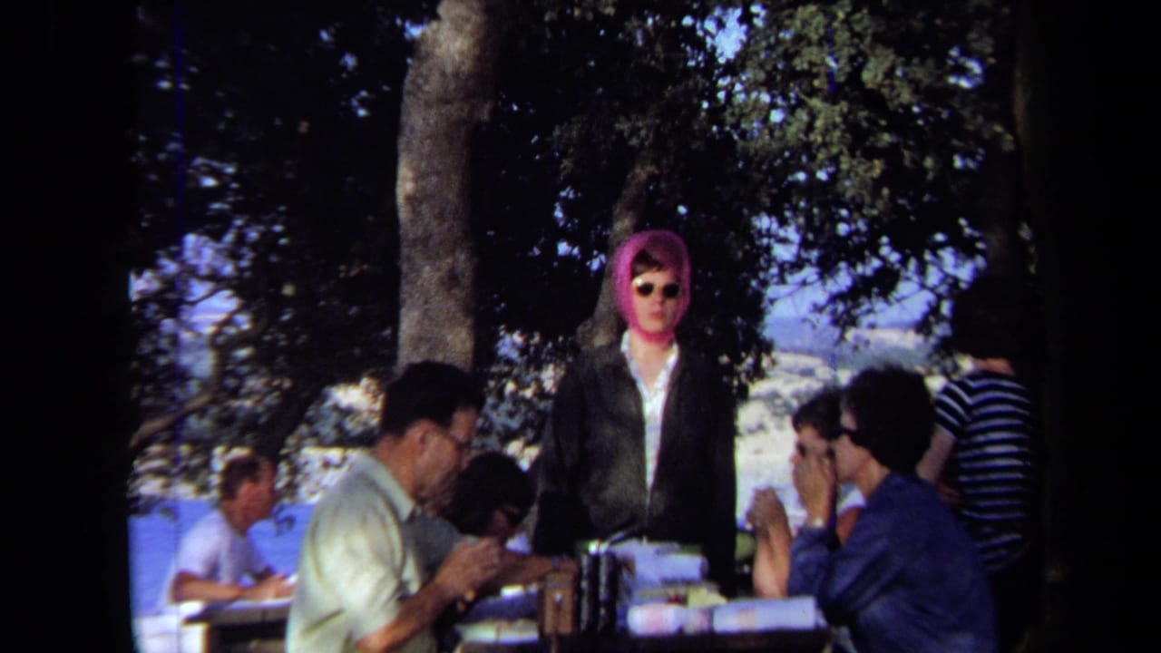 1971 California 8mm frame showing adults at a shaded outdoor picnic table with a woman in a pink headscarf and sunglasses standing at center.