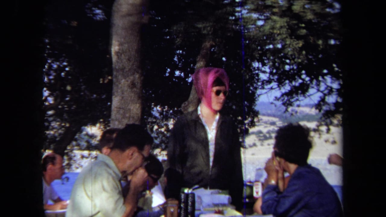 1971 California 8mm frame showing adults at a shaded picnic table near the ocean, including a standing figure in a vivid pink head covering and sunglasses.