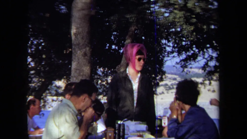 1971 California 8mm frame showing adults at a shaded picnic table near the ocean, including a standing figure in a vivid pink head covering and sunglasses.