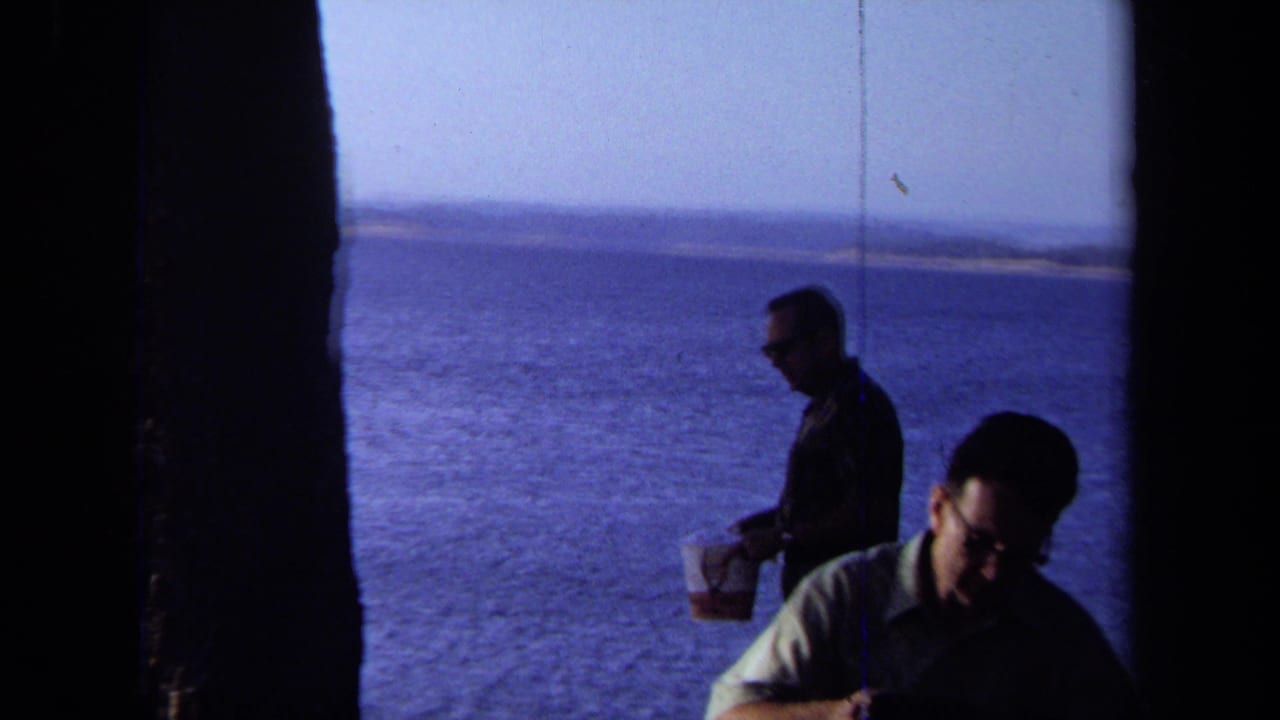 Two adults near the shoreline, one seated in shadow and one standing with a bucket.