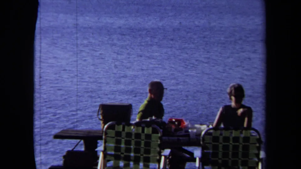 1971 California seaside picnic frame showing two adults seated in patterned folding chairs at a table overlooking blue water.