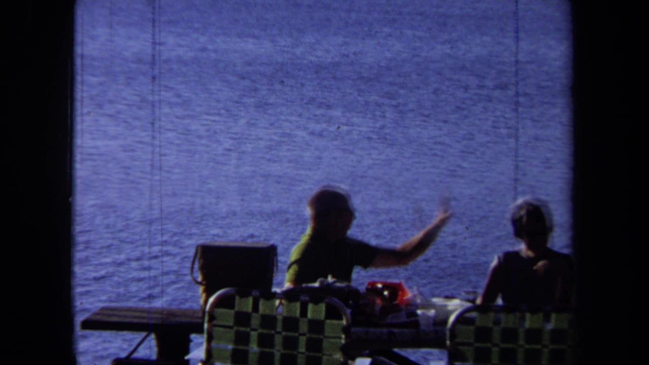 1971 California 8mm frame of two adults seated at a lakeside picnic table, one gesturing, with checkered chairs and blue water in the background.