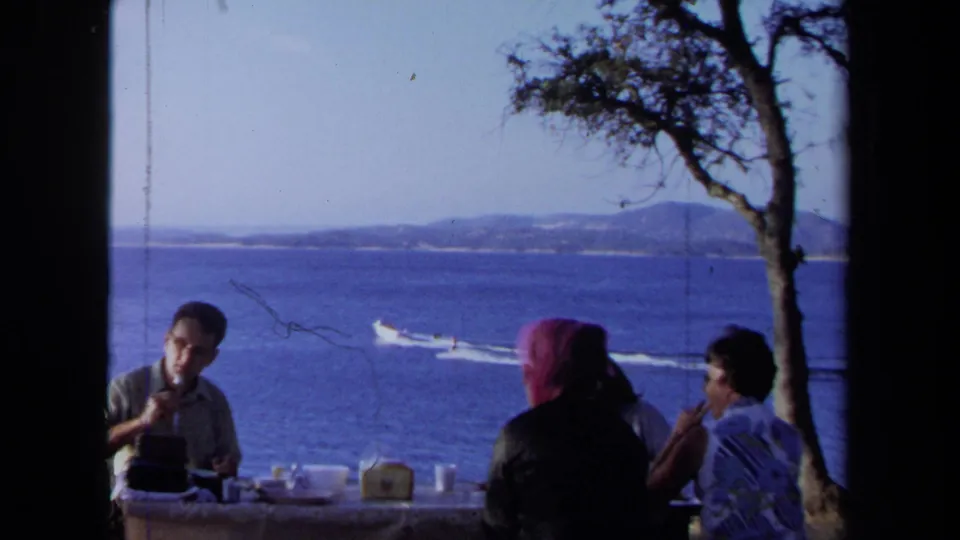 1971 California 8mm frame with adults at a lakeside picnic table, a tree at center, distant shoreline, and a speedboat wake on the water.