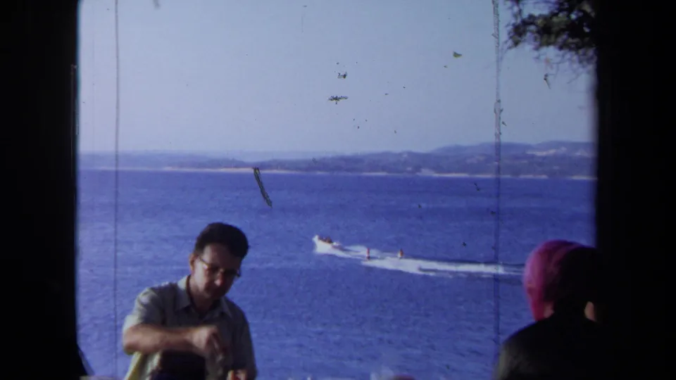 1971 California lake picnic frame with a seated man in the foreground, another person partly visible, and a speedboat leaving a white wake behind them.