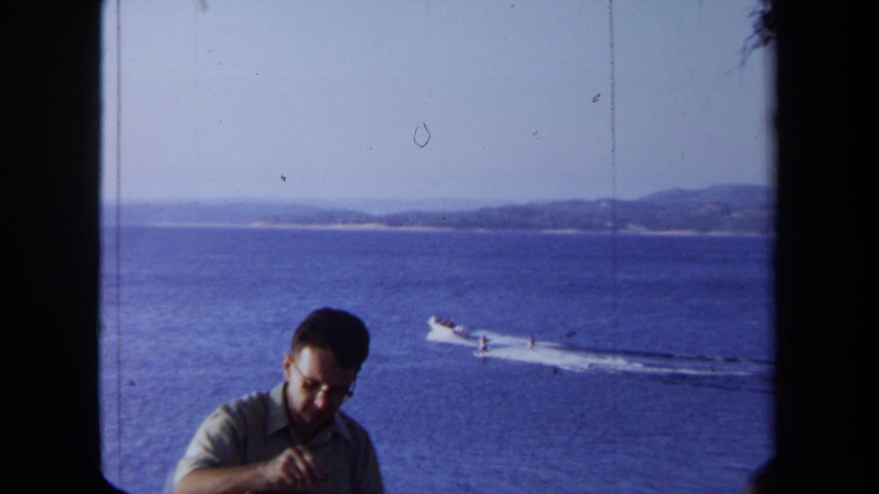 1971 California 8mm frame of a man in glasses near a window as a boat pulls a water skier across blue coastal water.
