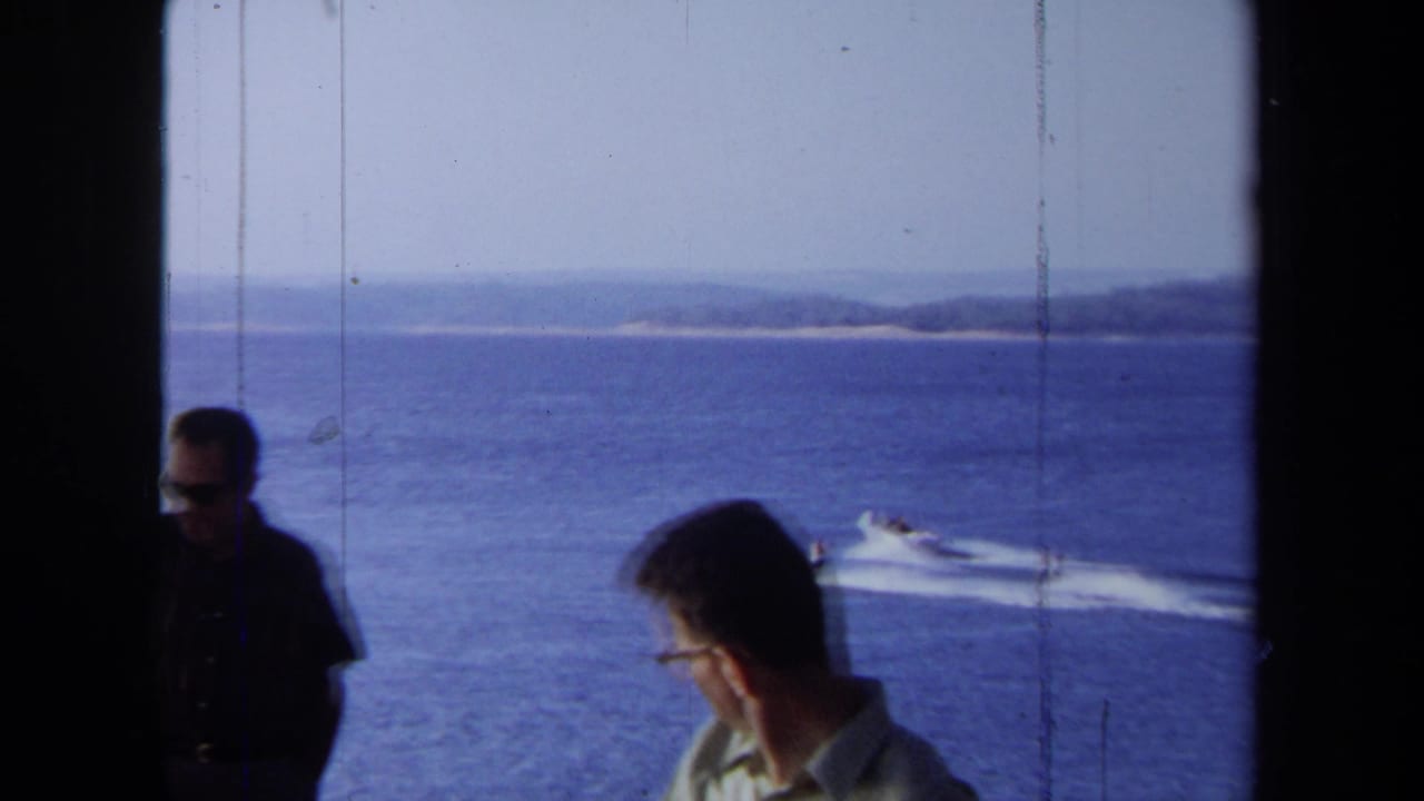 Two men near a window look toward the ocean as boats and wakes cross the water.