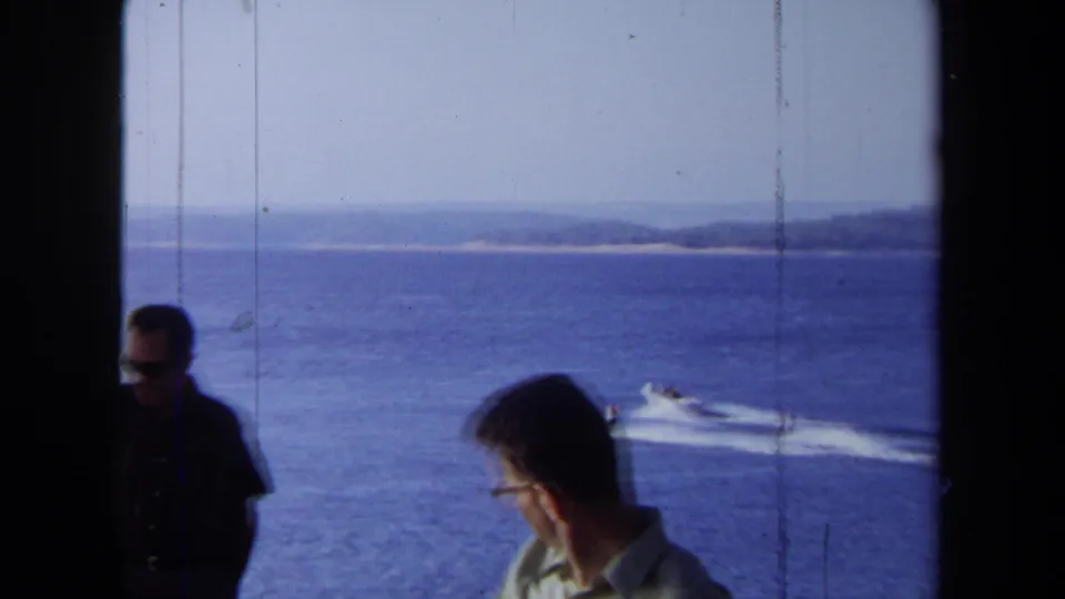 1971 California coastal home-movie frame with two adult men near a window, one in sunglasses, overlooking boats and water-ski wakes.