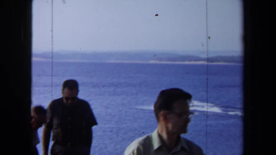 Home-movie frame from 1971 California showing adult men near an ocean overlook with a boat wake and distant shoreline visible.