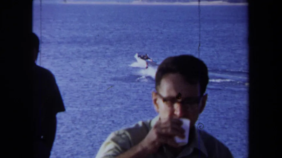 1971 California 8mm frame of a man in glasses drinking near the camera with a boat and water skier visible on the blue ocean.