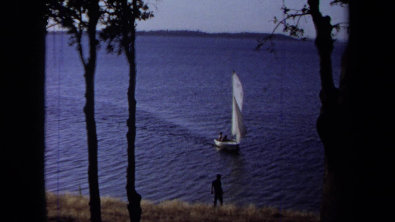1971 California 8mm frame of a white sailboat moving across blue lake water as a shoreline figure watches between tree silhouettes.