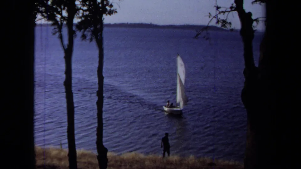 1971 California 8mm frame of a white sailboat moving across blue lake water as a shoreline figure watches between tree silhouettes.