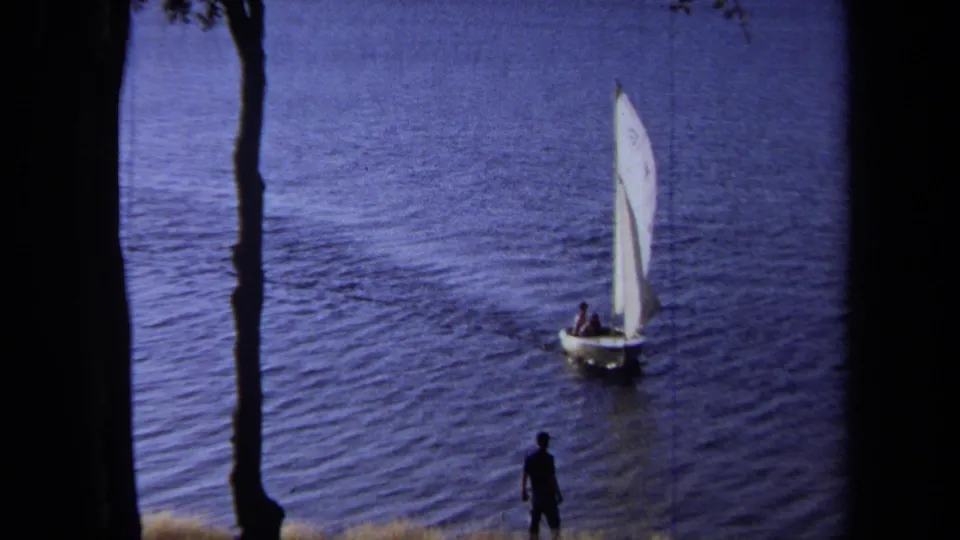 White-sailed dinghy glides across lake water in 1971 California home-movie footage, viewed through dark shoreline trees.
