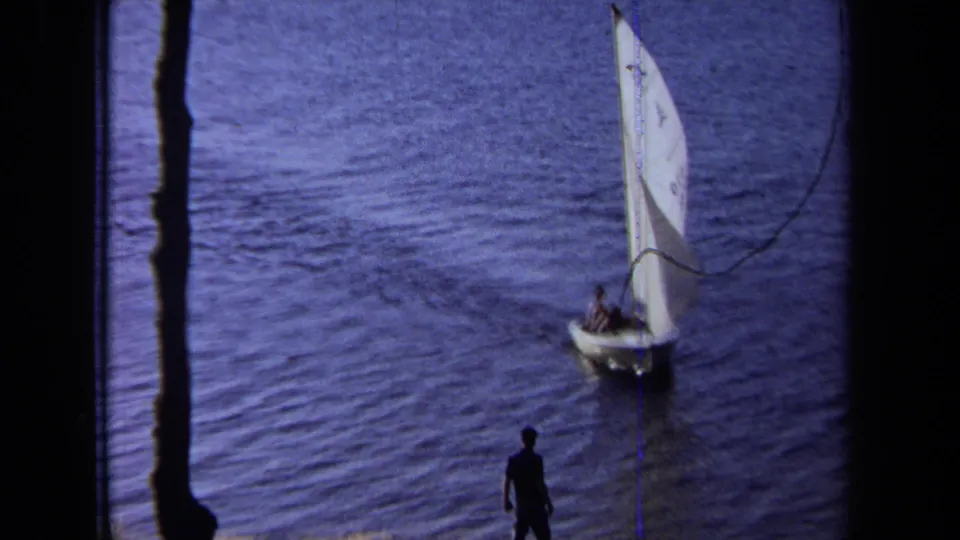 Closer 8mm view of a small sailboat near the wooded shoreline, with two occupants aboard and a silhouetted watcher below.