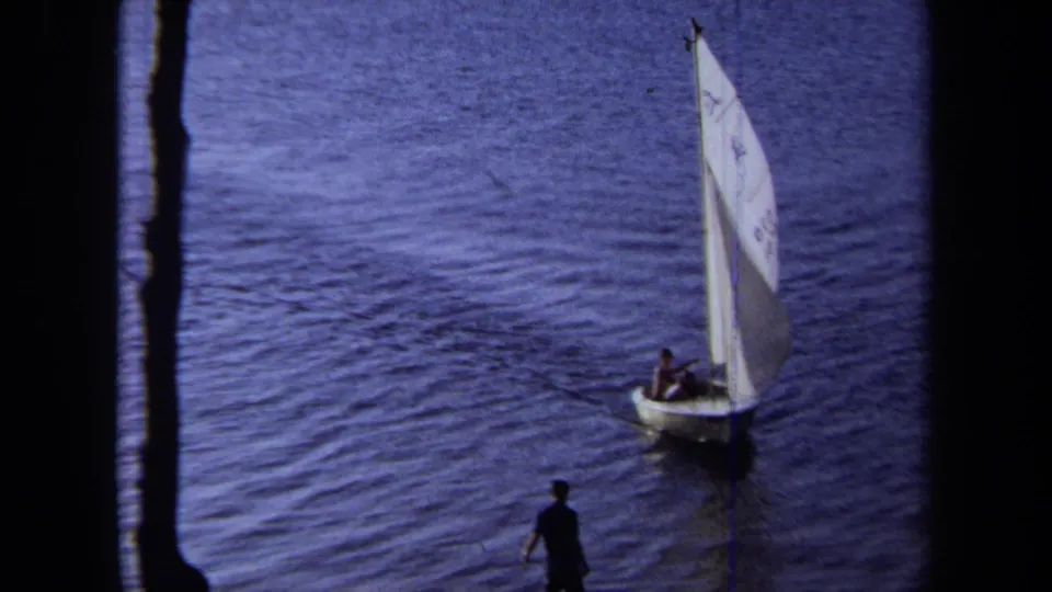 1971 California lake sailing frame with a small white sailboat and two distant people aboard, watched from a shaded bank.