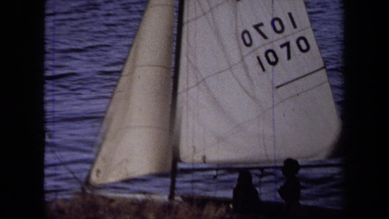 Small sailboat with a white sail marked 1070 near silhouetted figures by the water.