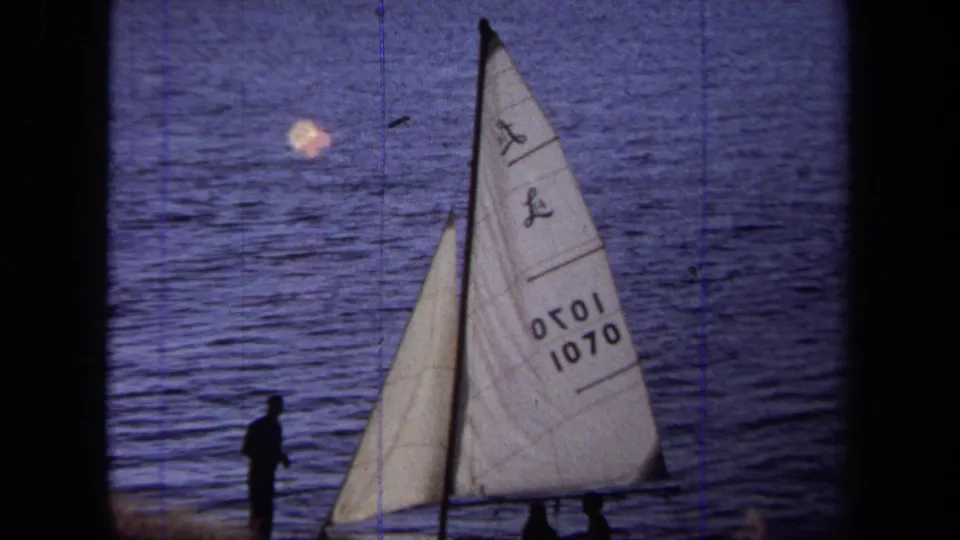 1971 California home-movie frame showing a white-sailed boat near shore, with a silhouetted man and calm blue water behind it.