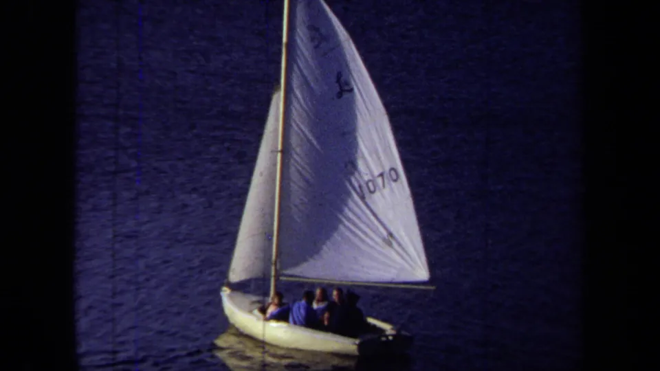1971 California 8mm frame showing a small sailboat on dark blue water with several seated passengers and a marked white sail.