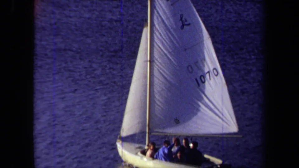 A closer 8mm view shows the small sailboat, its mast and numbered sail, and passengers seated low in the hull.