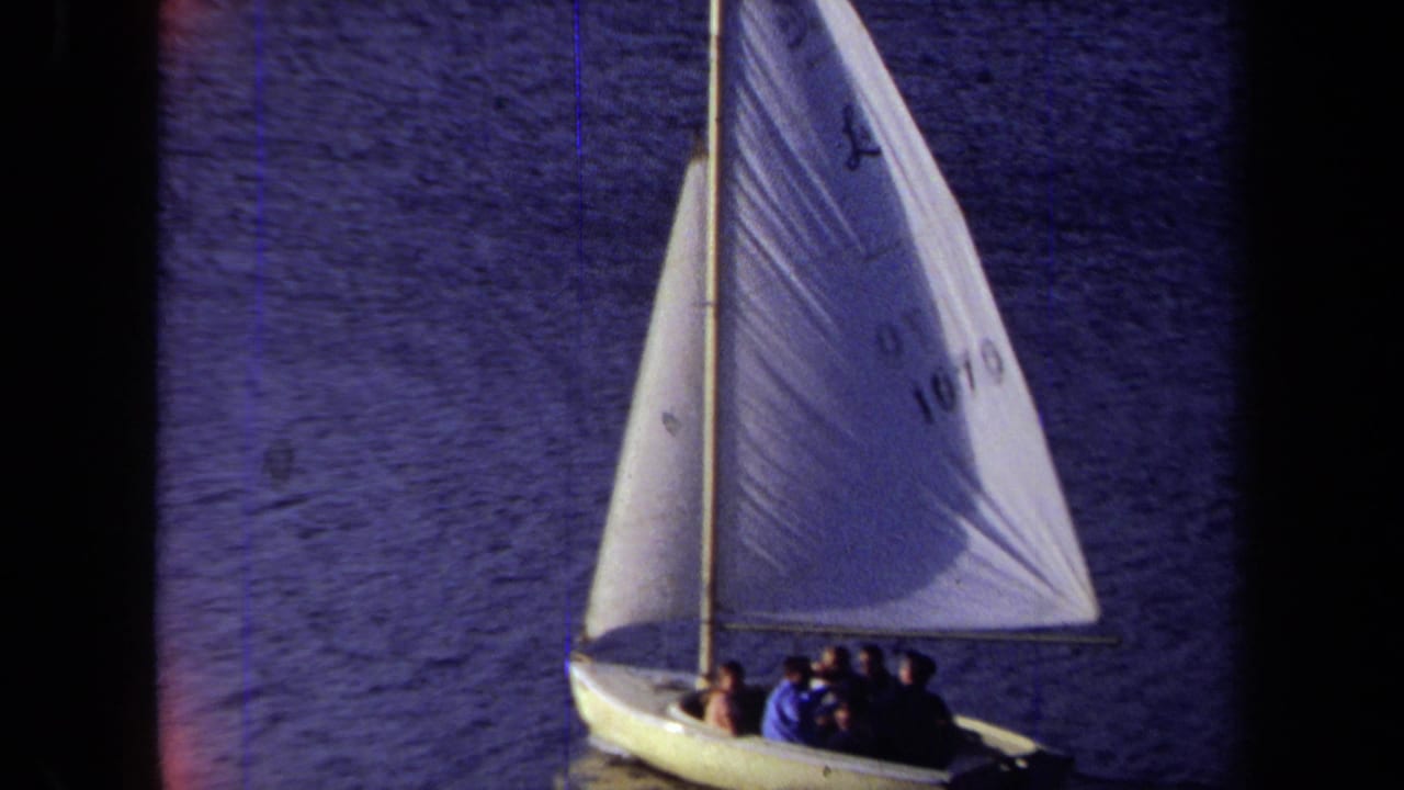 Small sailboat with a white sail carrying several people on dark blue water.