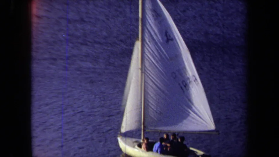 1971 California home-movie frame of a small sailboat and grouped passengers under a white sail with visible markings.