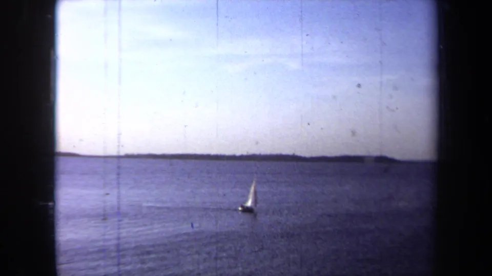 Wide 1971 California 8mm view of a small sailboat far out on calm water with a low shoreline in the distance.