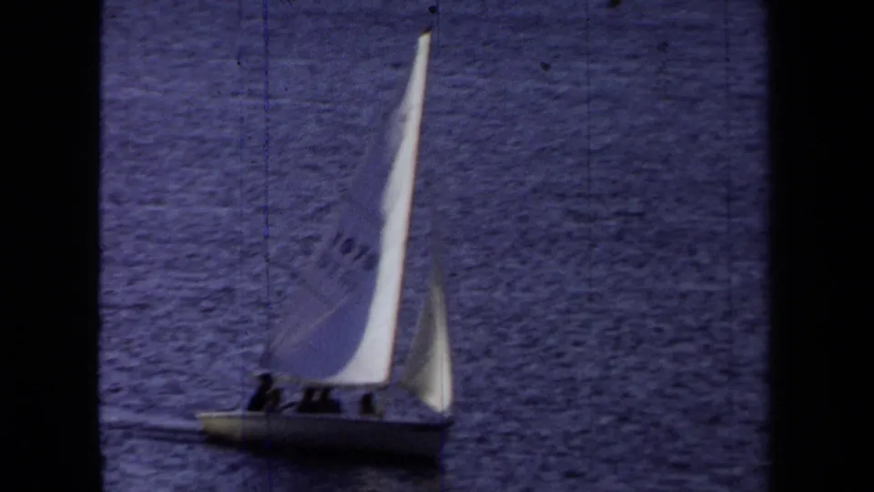 1971 California frame showing the small sailboat from farther away, with people aboard and sails angled over dark water.