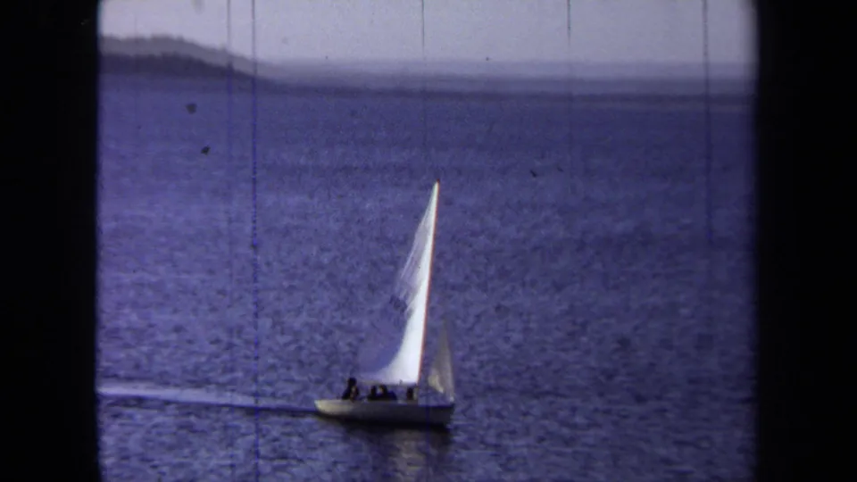 1971 California coastal frame with a small white sailboat carrying passengers under a soft overcast sky.