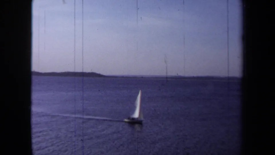 Wide 1971 California 8mm view of a sailboat receding across open coastal water toward a low shoreline.