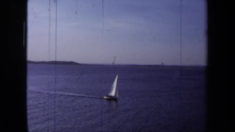 1971 California home-movie frame showing a distant white sailboat on calm blue water with a low shoreline beyond.