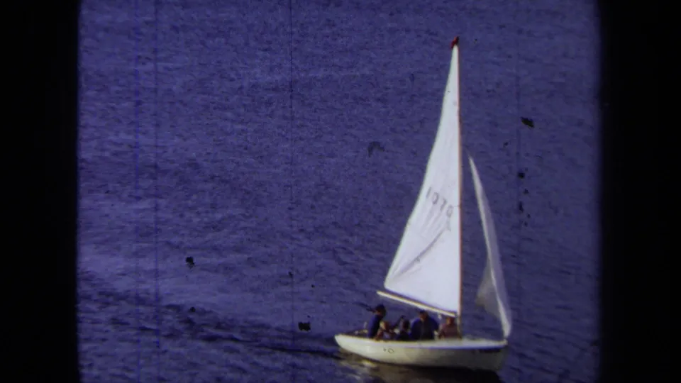 1971 California home-movie frame showing sail number 1070 and several people seated in a small white boat.