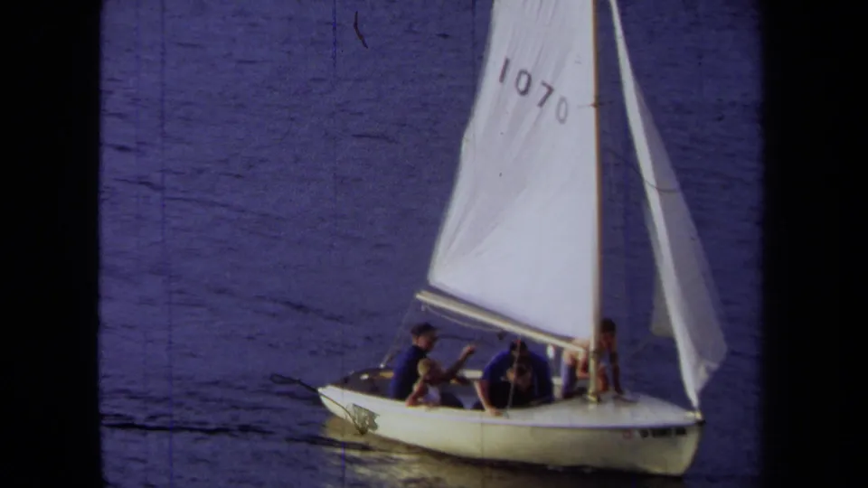 1971 California home-movie frame showing a closer view of passengers in a small white sailboat on blue water.