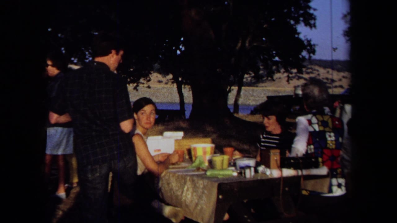 1971 California 8mm frame showing adults at a picnic table under trees, with lake water and dry hills behind them.
