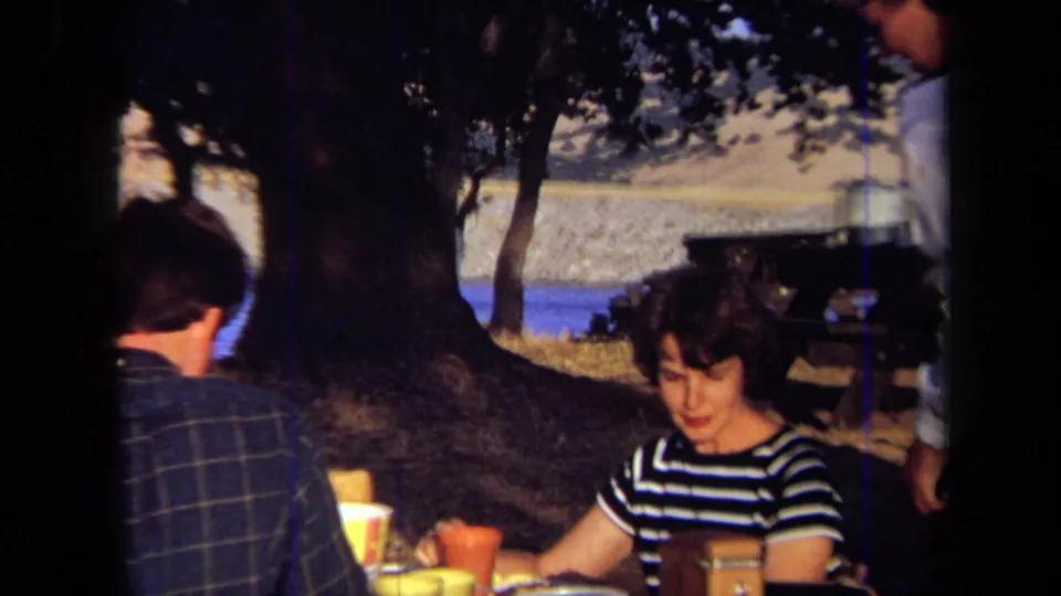 A woman in a striped shirt sits at the picnic table while another adult stands nearby, with lake and trees in the background.