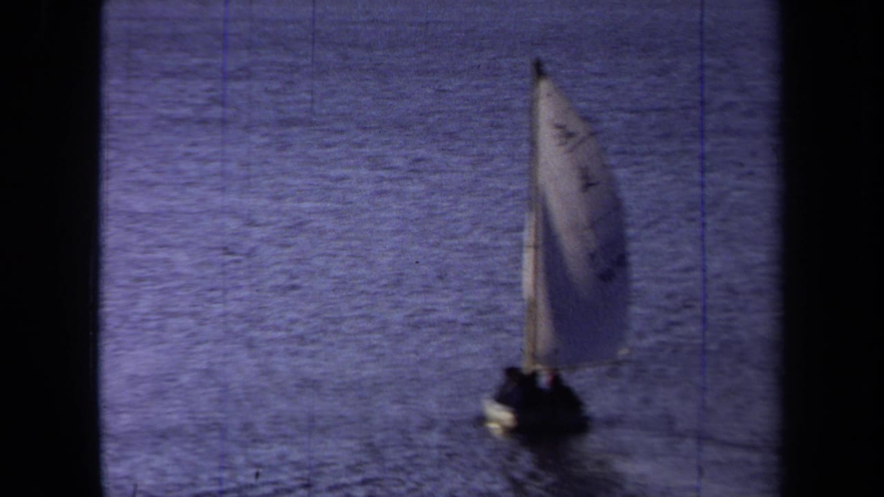 Sailboat farther across the water, its pale sail standing against the blue surface.